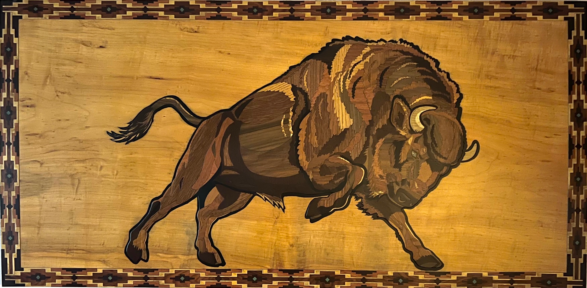 Wide wood inlay panel of a charging bison framed by a Southwestern border